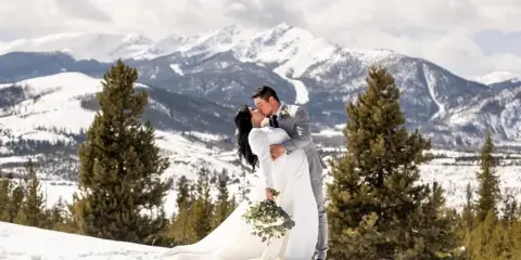 A bride and groom sharing a kiss on a snowy mountain slope with evergreen trees and snow-capped peaks in the background.