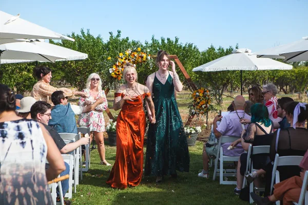 Two brides in colorful dresses walking down the aisle at an outdoor wedding ceremony.