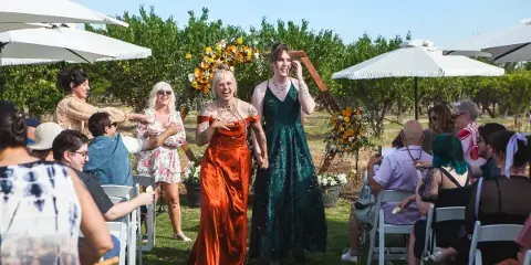 Two brides in colorful dresses walking down the aisle at an outdoor wedding ceremony.