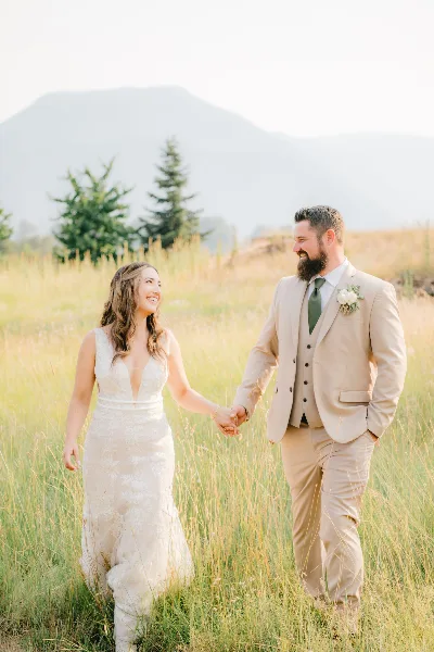 A bride and groom holding hands and walking through a grassy field with mountains in the background.