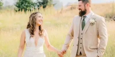 A bride and groom holding hands and walking through a grassy field with mountains in the background.