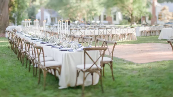An outdoor wedding reception setup featuring long tables with white linens, cross-back wooden chairs, and tall candle centerpieces.