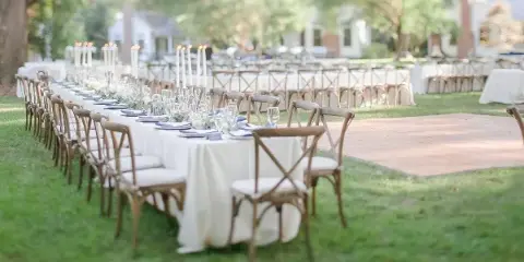 An outdoor wedding reception setup featuring long tables with white linens, cross-back wooden chairs, and tall candle centerpieces.