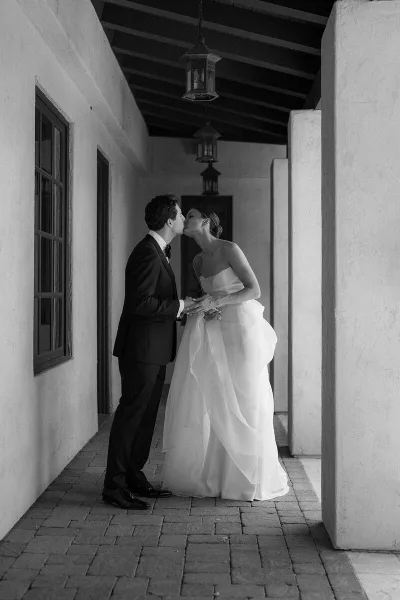 Black and white photograph of a bride and groom kissing in a corridor with hanging lanterns.