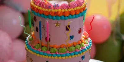 A colorful, two-tiered custom cake decorated with piped frosting, sprinkles, and candles on a wooden stool.