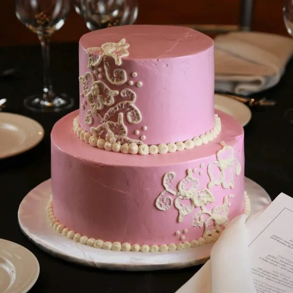 A two-tier pink wedding cake with white piped floral decorations and pearl borders on a white cake stand.