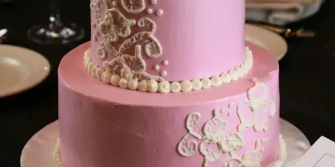A two-tier pink wedding cake with white piped floral decorations and pearl borders on a white cake stand.