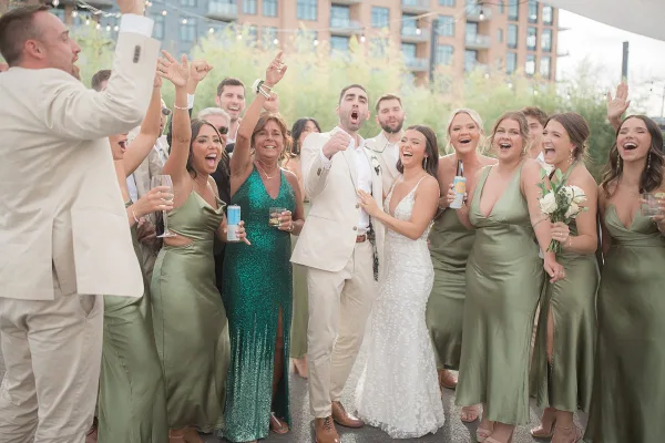 A bride and groom surrounded by their wedding party, all laughing and cheering with raised hands and drinks in an outdoor setting.