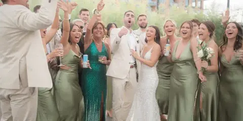 A bride and groom surrounded by their wedding party, all laughing and cheering with raised hands and drinks in an outdoor setting.