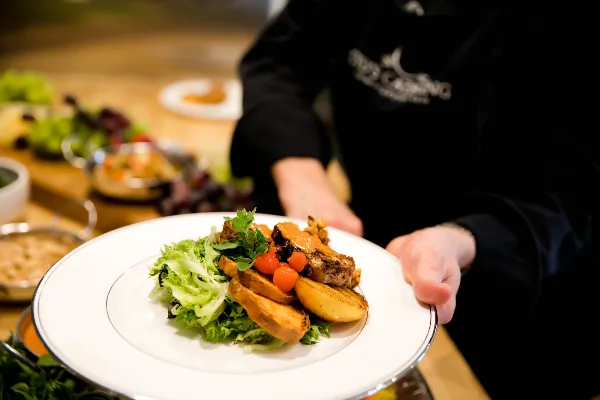 A catering server in a black uniform holding a white plate with a plated meal featuring meat, potatoes, and greens.