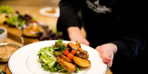 A catering server in a black uniform holding a white plate with a plated meal featuring meat, potatoes, and greens.