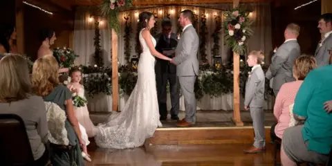 A bride and groom exchanging vows during a wedding ceremony under a decorated wooden arbor in an indoor event venue.