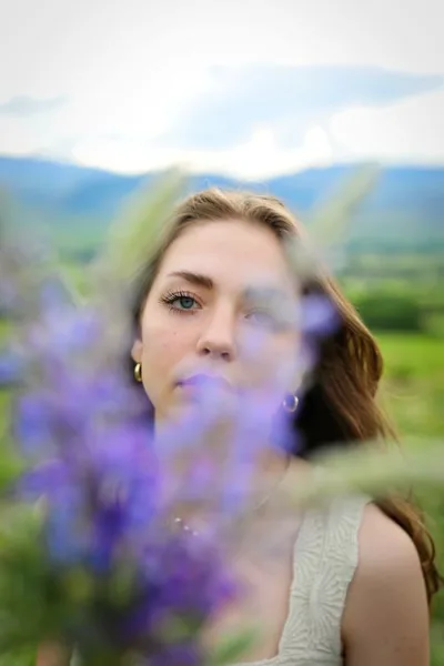 Outdoor portrait of a woman with blue eyes and brown hair, framed by blurred purple flowers in the foreground with a mountain landscape in the background.
