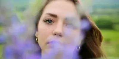 Outdoor portrait of a woman with blue eyes and brown hair, framed by blurred purple flowers in the foreground with a mountain landscape in the background.