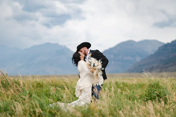A bride and groom kissing in a grassy field with mountains in the background.