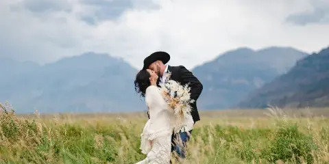 A bride and groom kissing in a grassy field with mountains in the background.