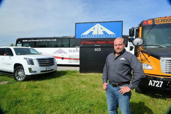 A man stands in front of an AA Transportation sign, a white limousine, and a yellow school bus.