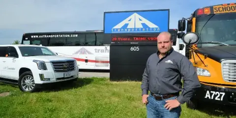 A man stands in front of an AA Transportation sign, a white limousine, and a yellow school bus.