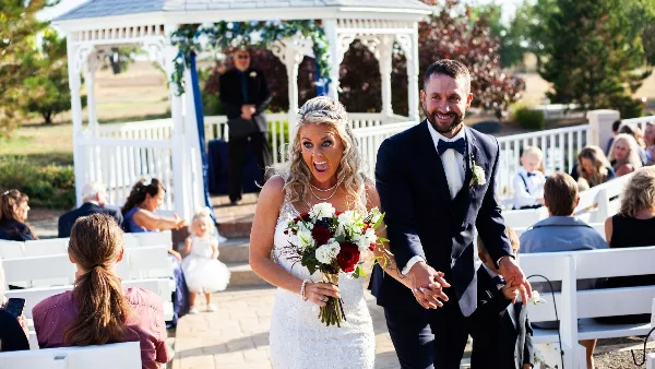 A bride and groom walking down the aisle at an outdoor wedding ceremony with a white gazebo in the background.