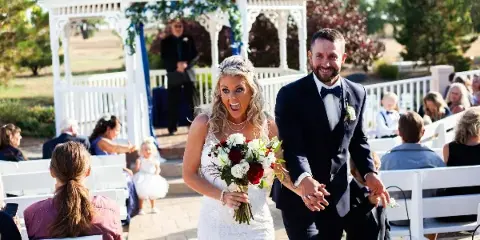 A bride and groom walking down the aisle at an outdoor wedding ceremony with a white gazebo in the background.