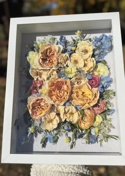 A framed pressed flower arrangement featuring preserved roses and blue flowers.