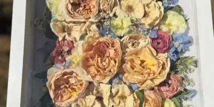 A framed pressed flower arrangement featuring preserved roses and blue flowers.