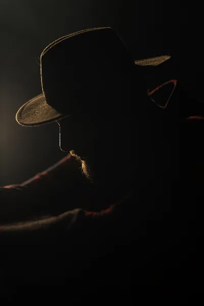 Dramatic low-key profile portrait of a bearded man wearing a hat, lit by a single rim light.