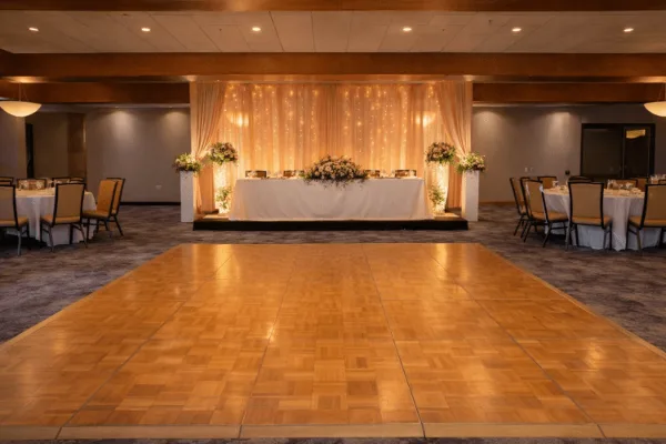Indoor event venue with a large wooden dance floor, round tables with white linens, and a decorated head table with floral arrangements.