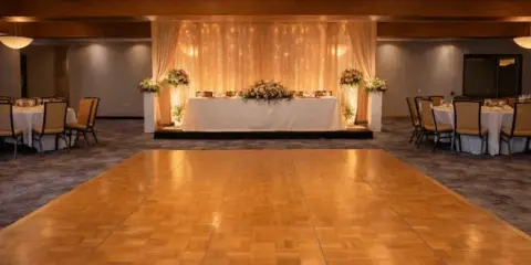 Indoor event venue with a large wooden dance floor, round tables with white linens, and a decorated head table with floral arrangements.