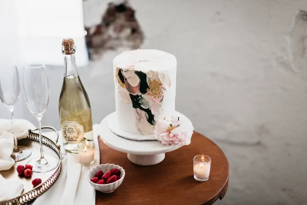 A white frosted cake with artistic gold and pink accents displayed on a white cake stand, accompanied by champagne and raspberries.