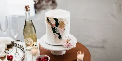 A white frosted cake with artistic gold and pink accents displayed on a white cake stand, accompanied by champagne and raspberries.