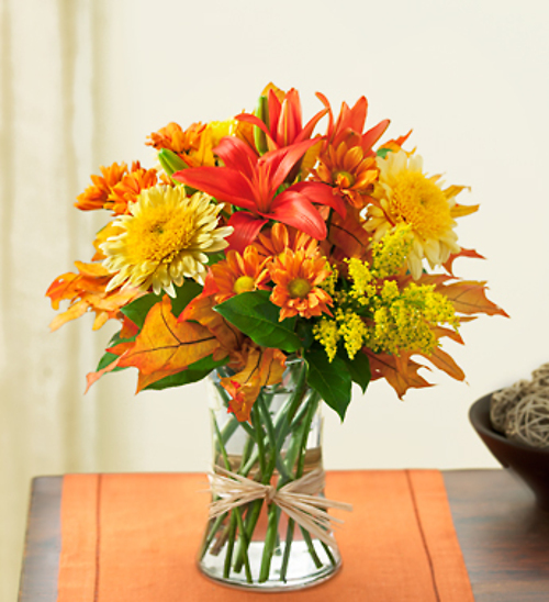 A fall-themed floral arrangement with orange lilies, yellow chrysanthemums, and autumn leaves in a clear glass vase.