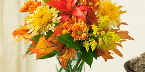 A fall-themed floral arrangement with orange lilies, yellow chrysanthemums, and autumn leaves in a clear glass vase.