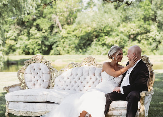 A bride and groom sitting together on an ornate white sofa in an outdoor setting near a body of water.