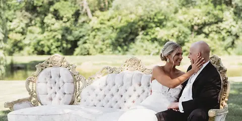 A bride and groom sitting together on an ornate white sofa in an outdoor setting near a body of water.