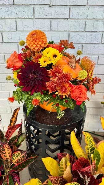 A vibrant fall-themed floral arrangement with orange dahlias, yellow chrysanthemums, and a decorative pumpkin displayed on a black stand.
