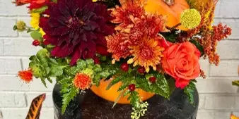 A vibrant fall-themed floral arrangement with orange dahlias, yellow chrysanthemums, and a decorative pumpkin displayed on a black stand.