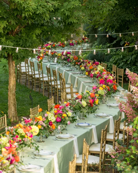 An outdoor event table set with green and white linens, gold chiavari chairs, and vibrant floral centerpieces.