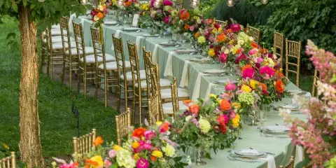 An outdoor event table set with green and white linens, gold chiavari chairs, and vibrant floral centerpieces.