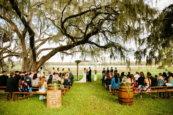 Outdoor wedding ceremony under a large oak tree with Spanish moss, overlooking a lake.