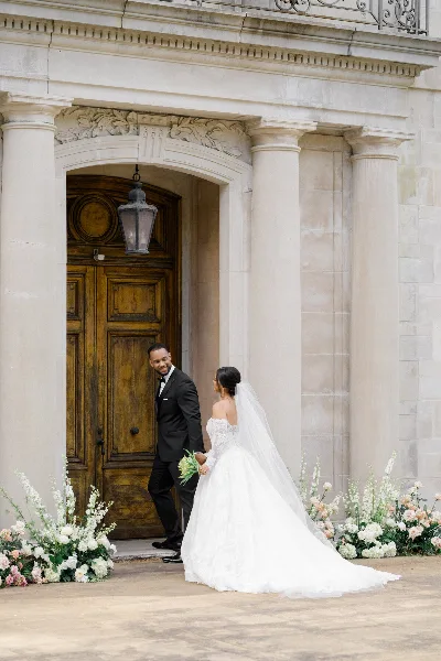 A bride in a white gown and a groom in a black tuxedo standing together in front of an ornate stone building entrance with floral arrangements.