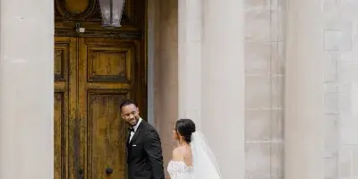 A bride in a white gown and a groom in a black tuxedo standing together in front of an ornate stone building entrance with floral arrangements.