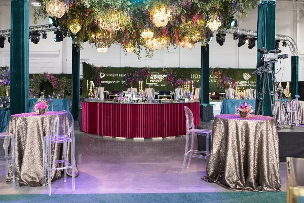 A professionally designed event venue featuring a lush floral ceiling installation, chandeliers, and a branded backdrop with corporate logos.