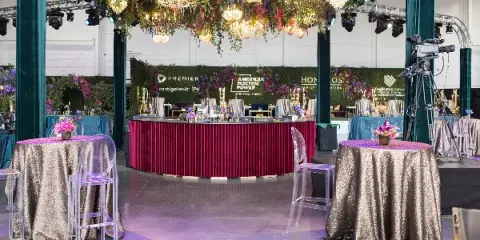 A professionally designed event venue featuring a lush floral ceiling installation, chandeliers, and a branded backdrop with corporate logos.
