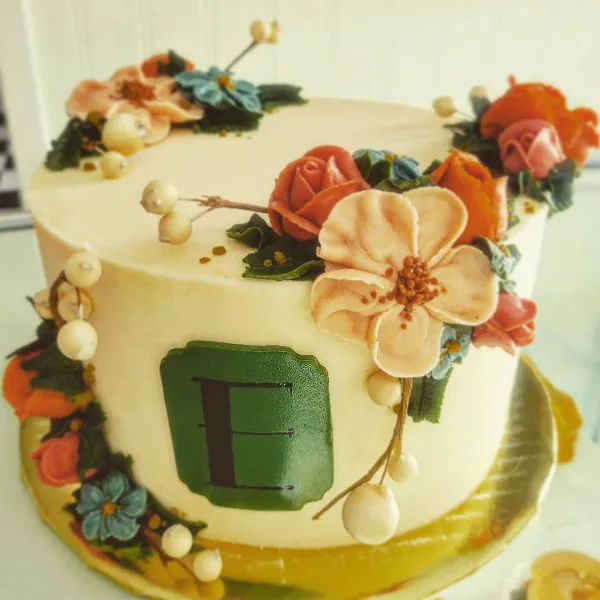 A close-up of a white frosted cake decorated with intricate buttercream flowers in shades of orange, pink, and blue, featuring a green monogrammed plaque on the side.
