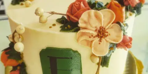 A close-up of a white frosted cake decorated with intricate buttercream flowers in shades of orange, pink, and blue, featuring a green monogrammed plaque on the side.
