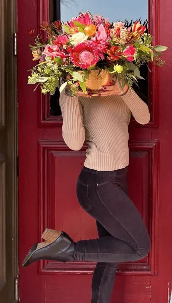 A person holding a large, colorful floral arrangement in front of a red door.