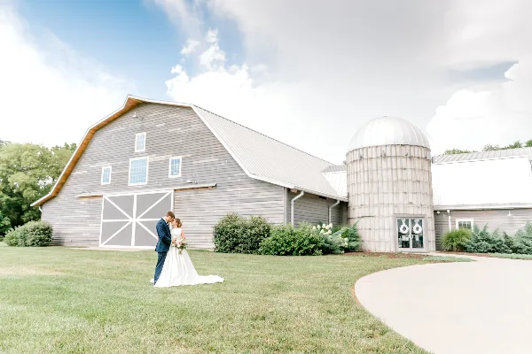A bride and groom standing on the lawn in front of a large grey barn-style building with a prominent silo structure.