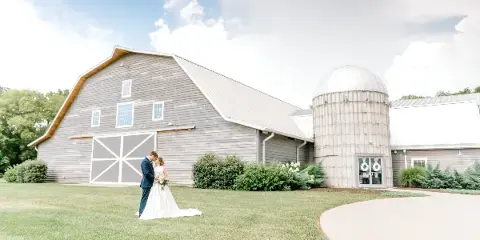 A bride and groom standing on the lawn in front of a large grey barn-style building with a prominent silo structure.