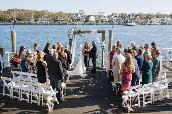 A wedding ceremony taking place on a dock with a wooden arch, white chairs, and floral arrangements.
